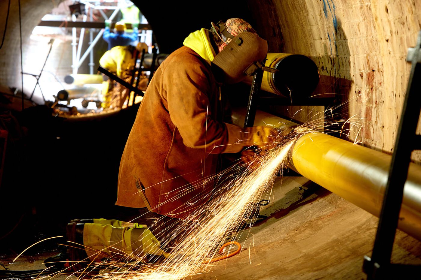 Foto: Bauarbeiter in Schutzausrüstung beim Schweißen einer Rohrleitung, rechts gewölbte Ziegelwand, unscharf im Hintergrund zwei weitere Arbeiter an einem Gerüst im Tageslicht